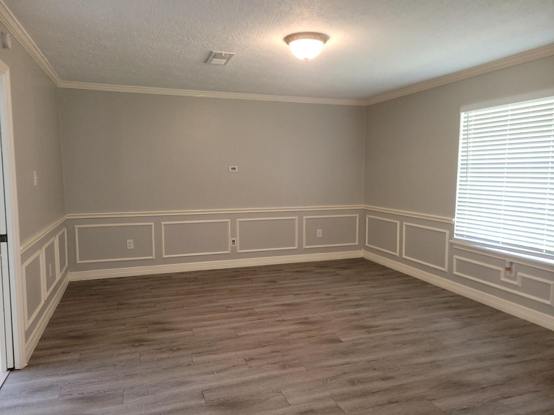 Empty room with grey walls, white trim, and wood-look flooring. Window with blinds.