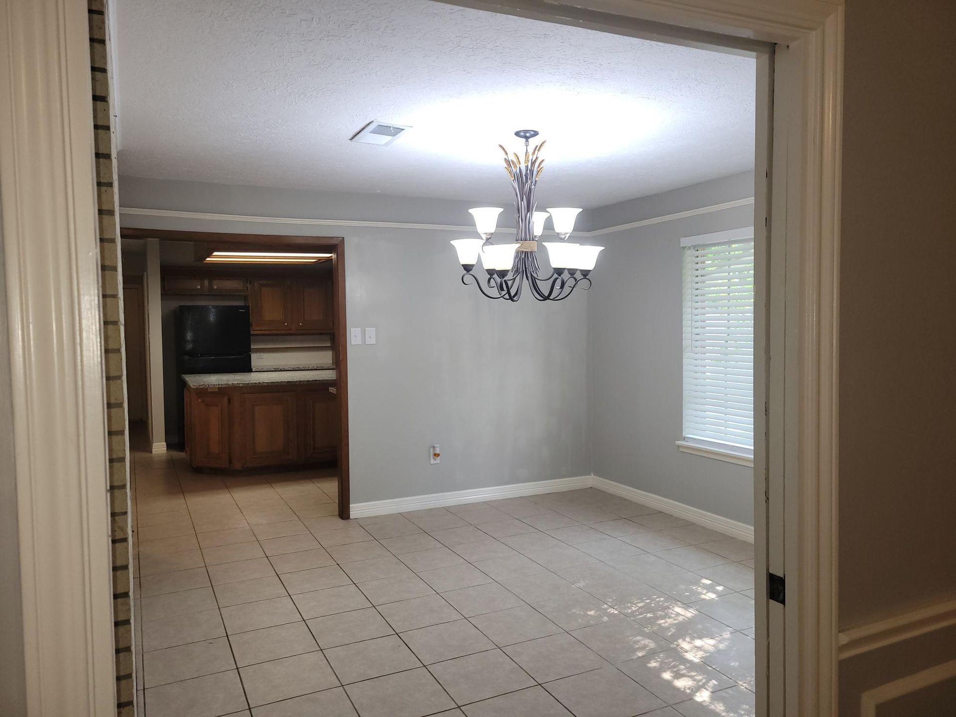 View of an empty dining room with chandelier, neutral walls, tiled floor, and a glimpse into a kitchen.