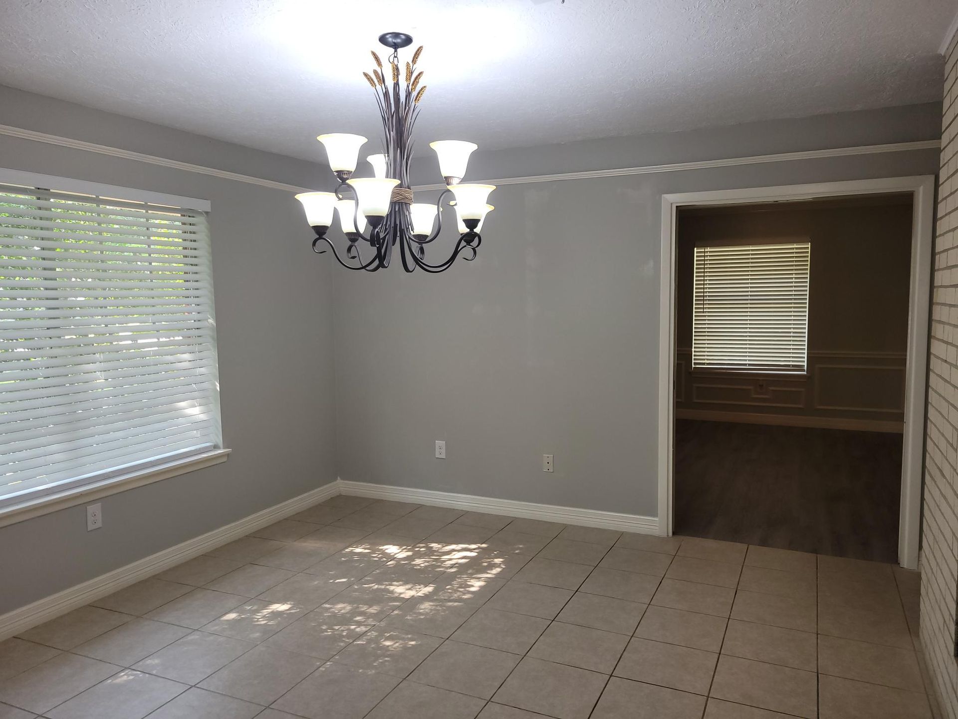 Empty dining room with chandelier, window with blinds, gray walls, doorway to another room.