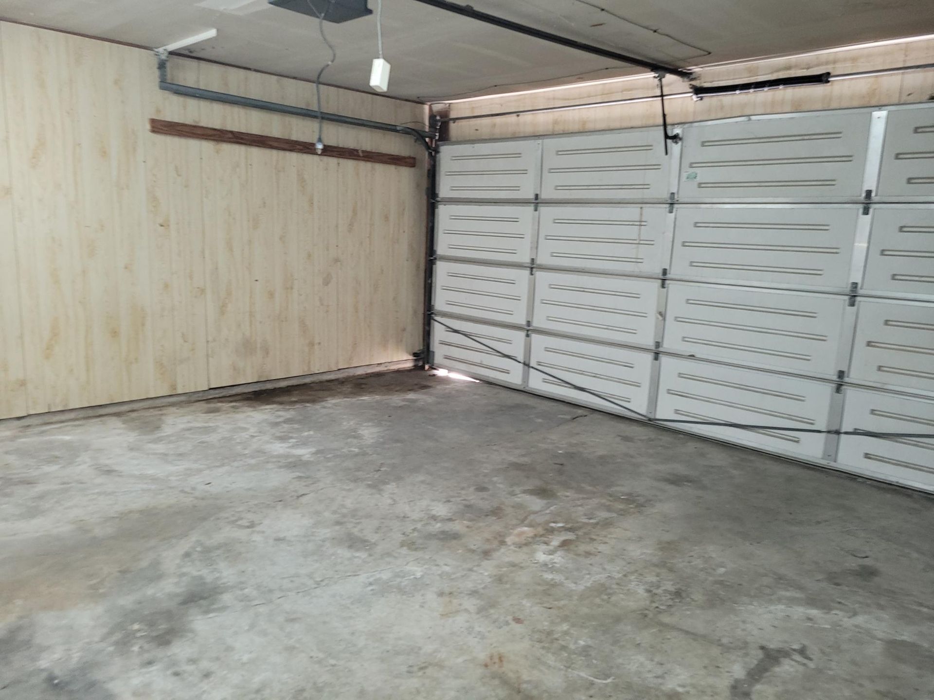 Empty garage with a concrete floor, a partially open white garage door, and light-colored wooden paneling on one wall.