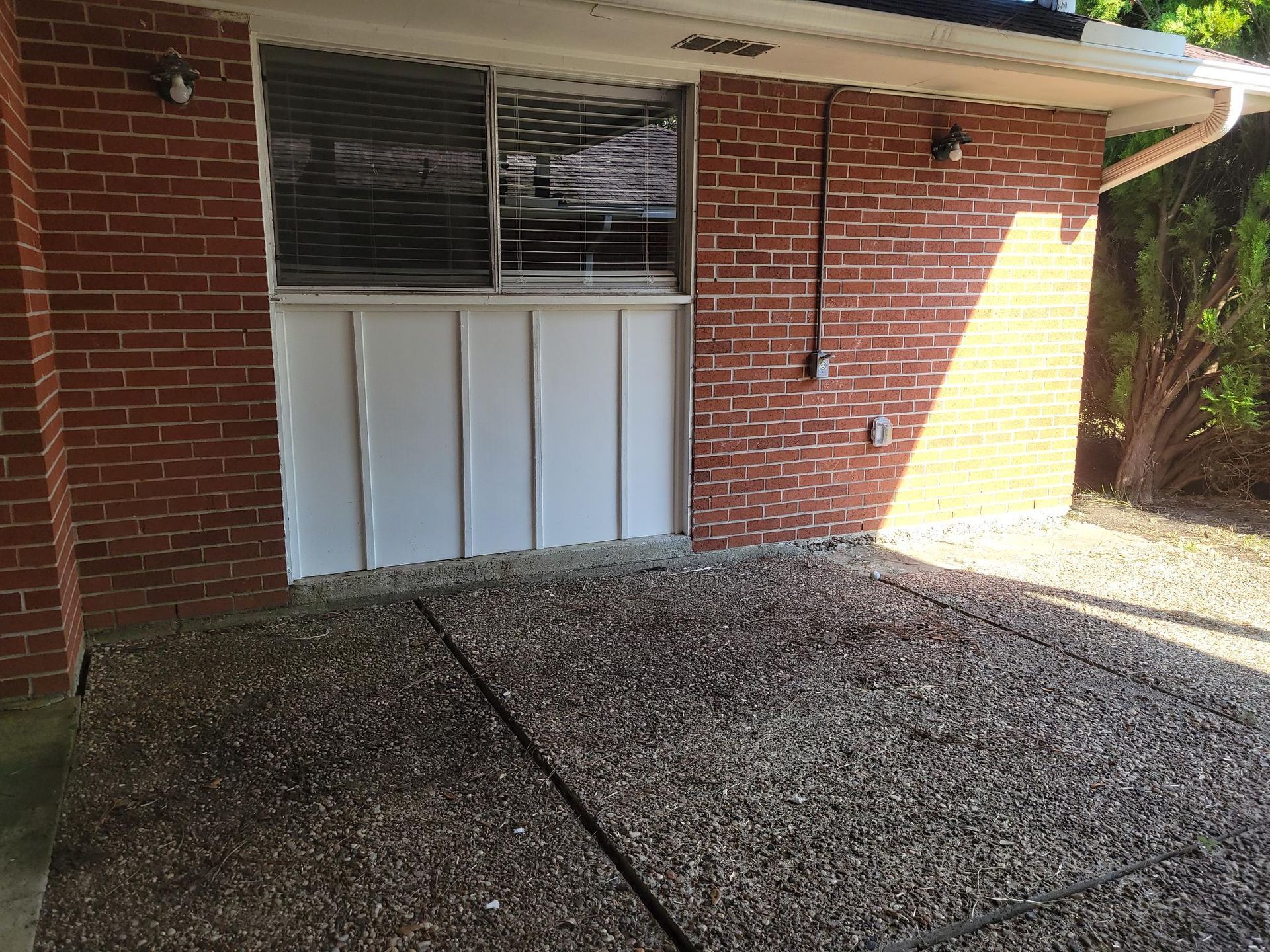 Exterior patio with brick walls, window with blinds, and pebbled floor.