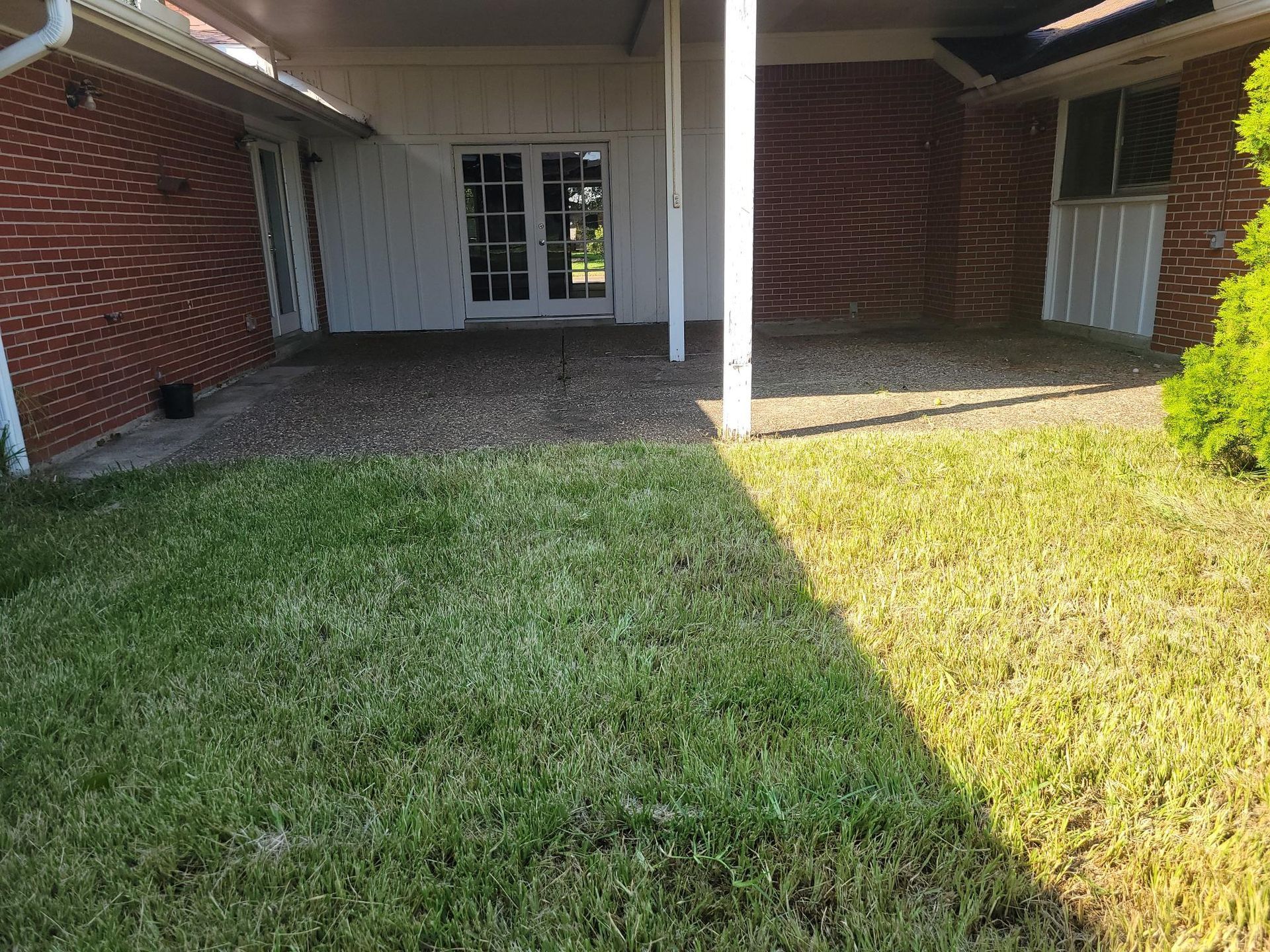 Green lawn in front of a shaded patio with a brick wall and white columns.