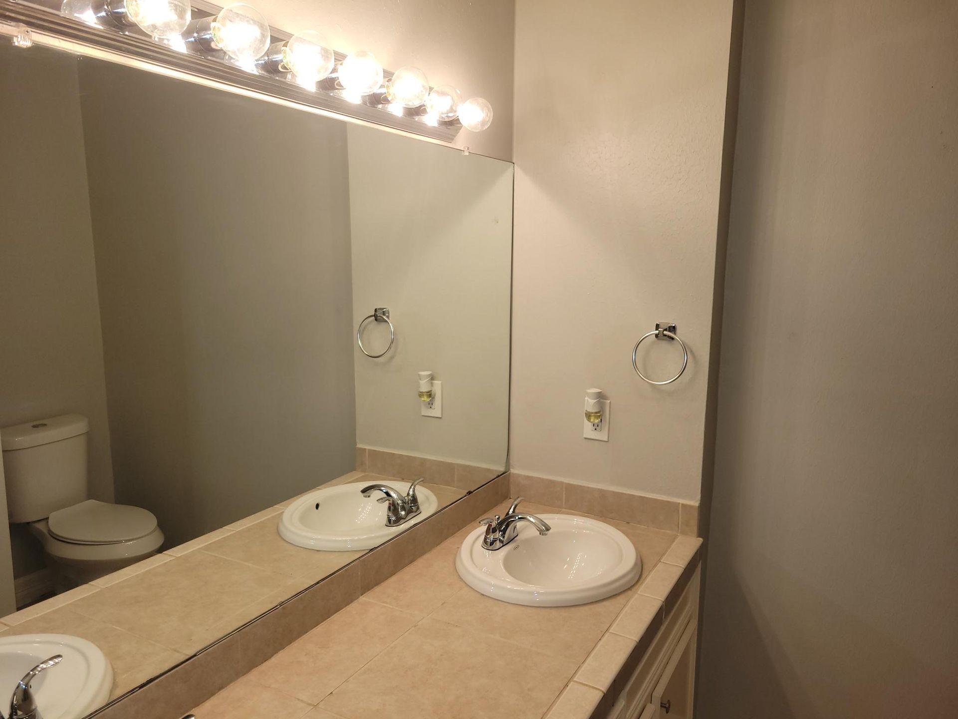 Bathroom with mirror, sinks, toilet, and overhead lights. Gray walls and beige countertop.