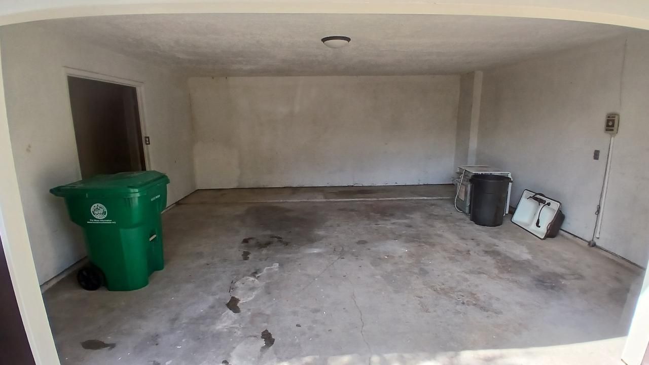 Empty garage with green trash bin, dirty concrete floor, and plain walls.