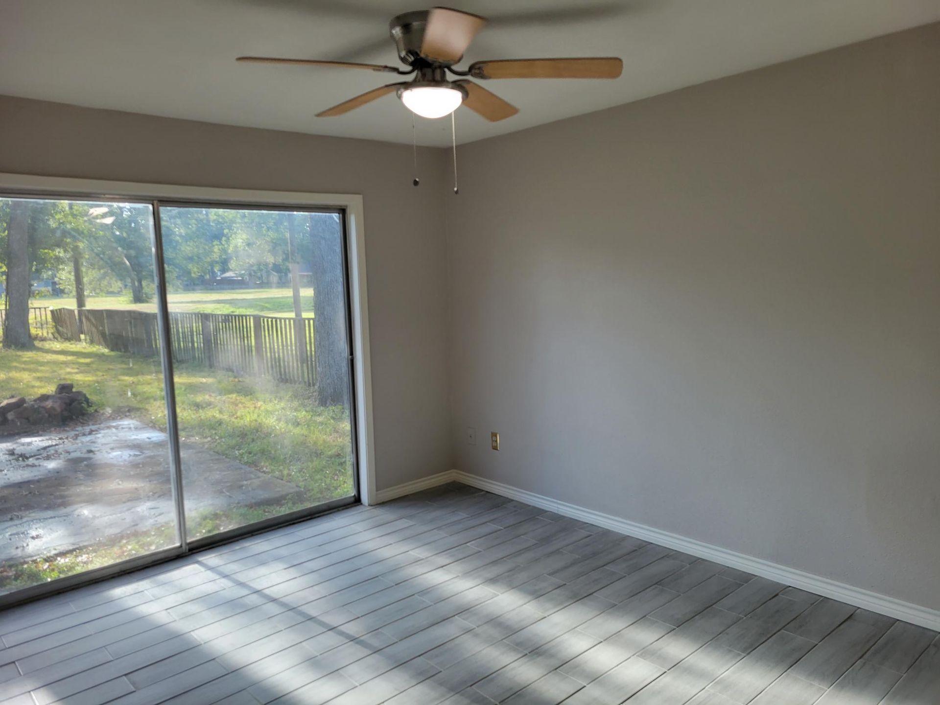 Empty room with gray walls and wood-look flooring. Sliding glass door leads to a yard. Ceiling fan.