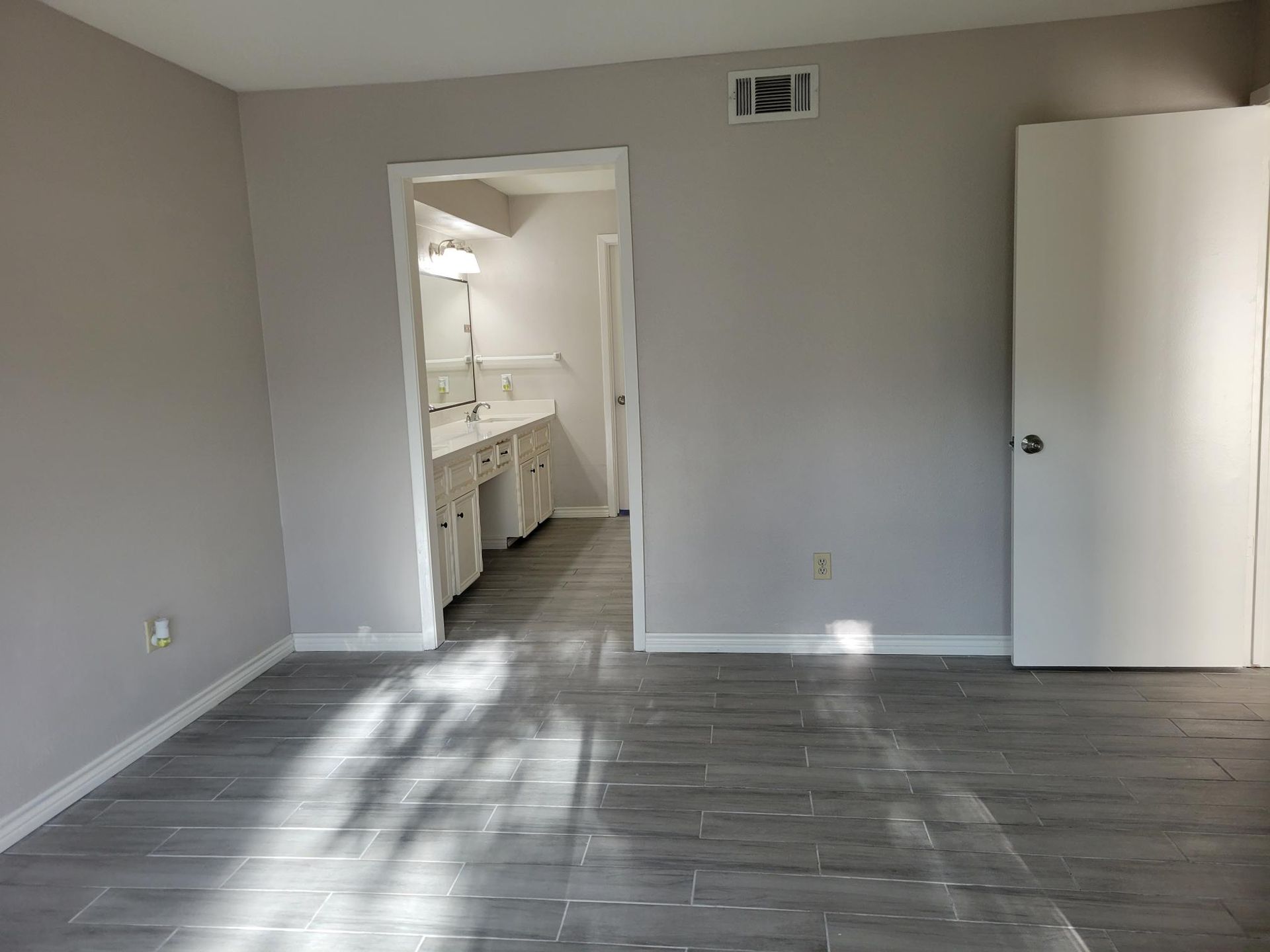 Empty bedroom with gray walls, wood-look flooring, and an open doorway to a bathroom.