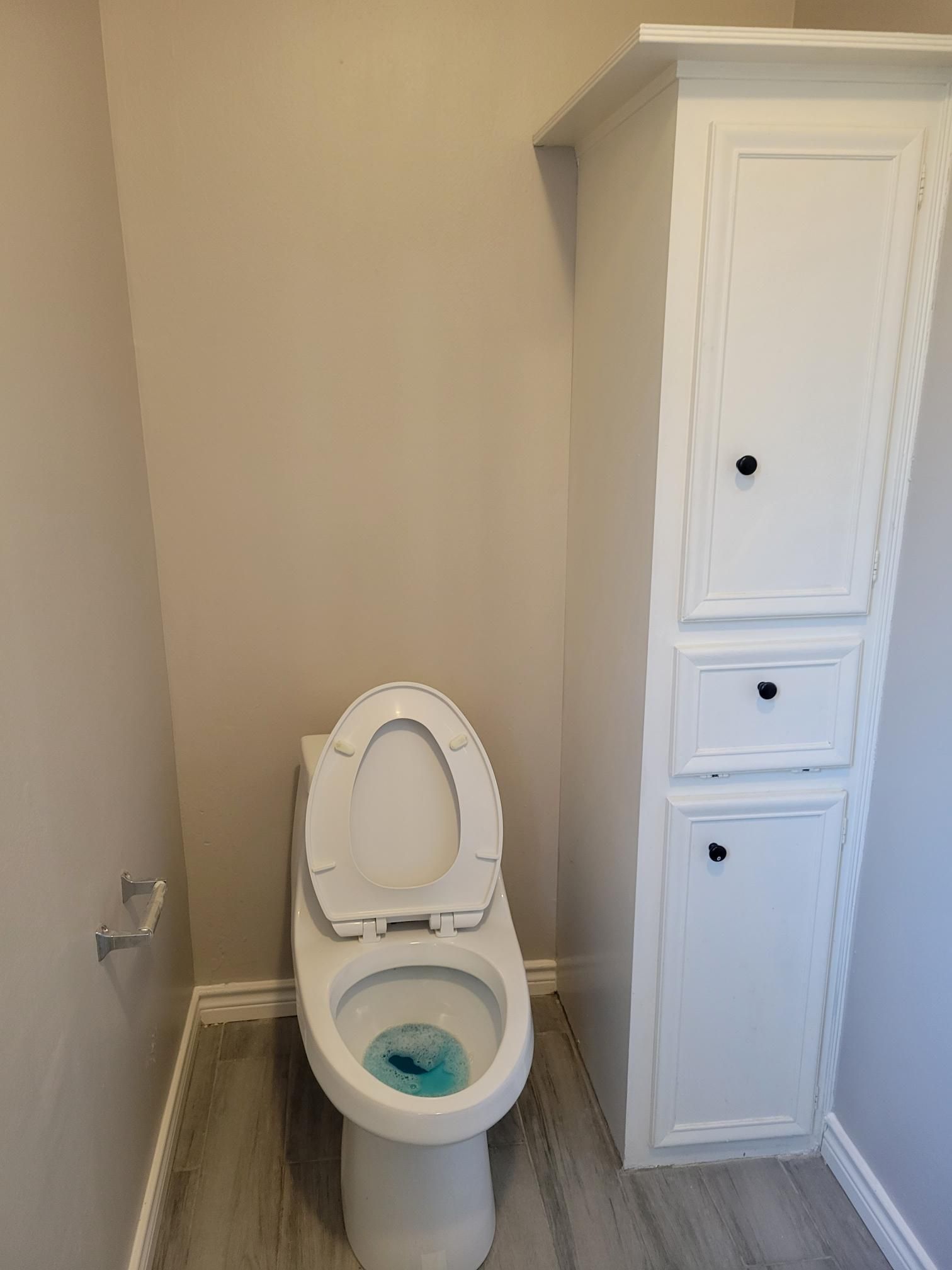 Toilet in bathroom with open lid, next to white storage cabinet. Gray walls and flooring.