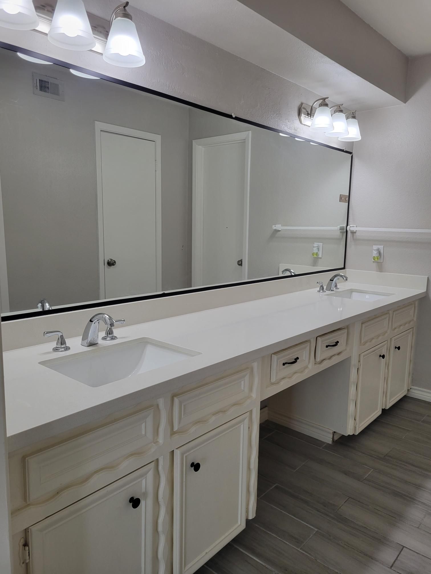 Bathroom with double sinks, large mirror, white cabinets, and gray flooring.