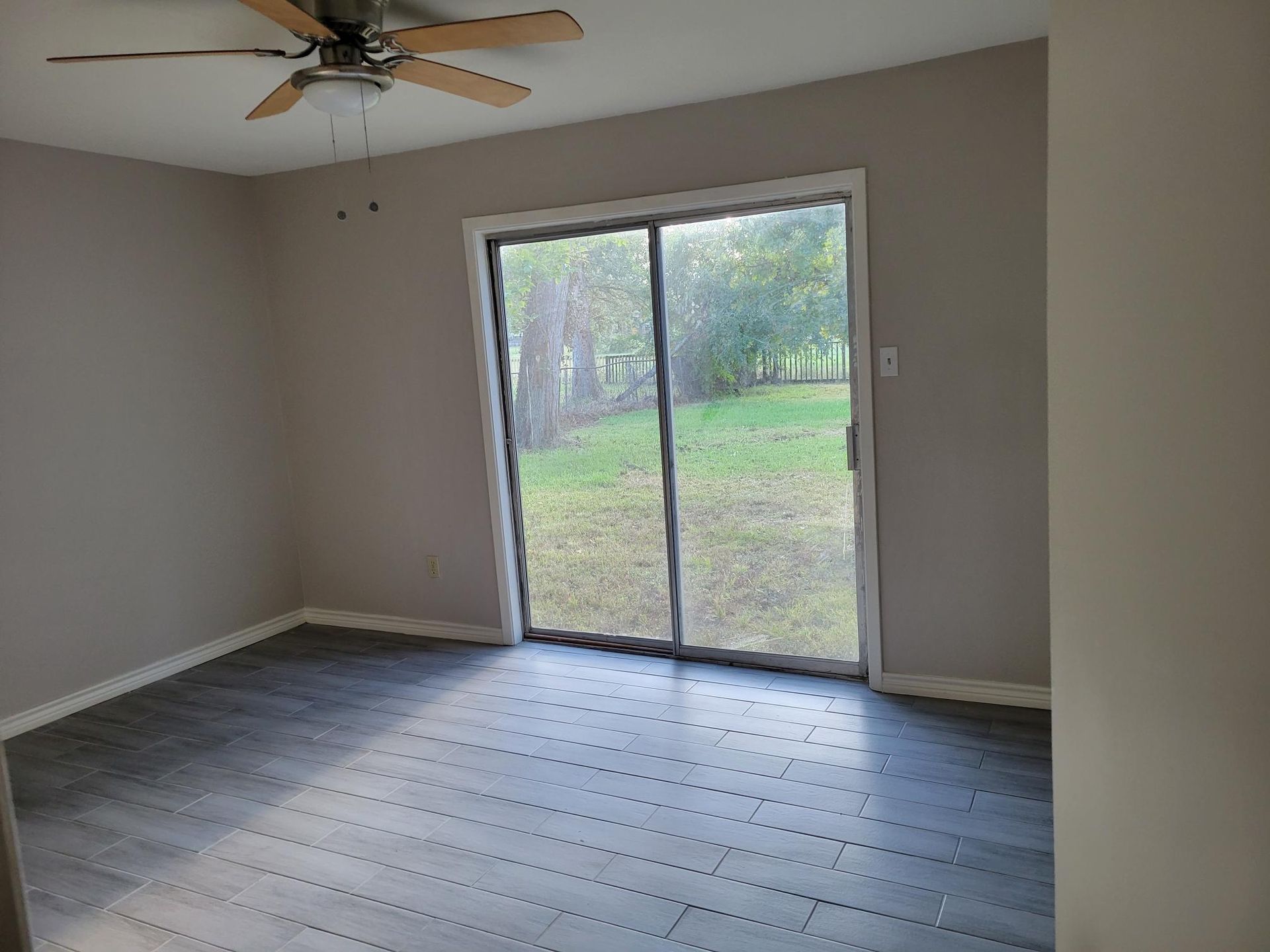 Empty room with gray floors and walls, sliding glass door to a grassy yard, ceiling fan.