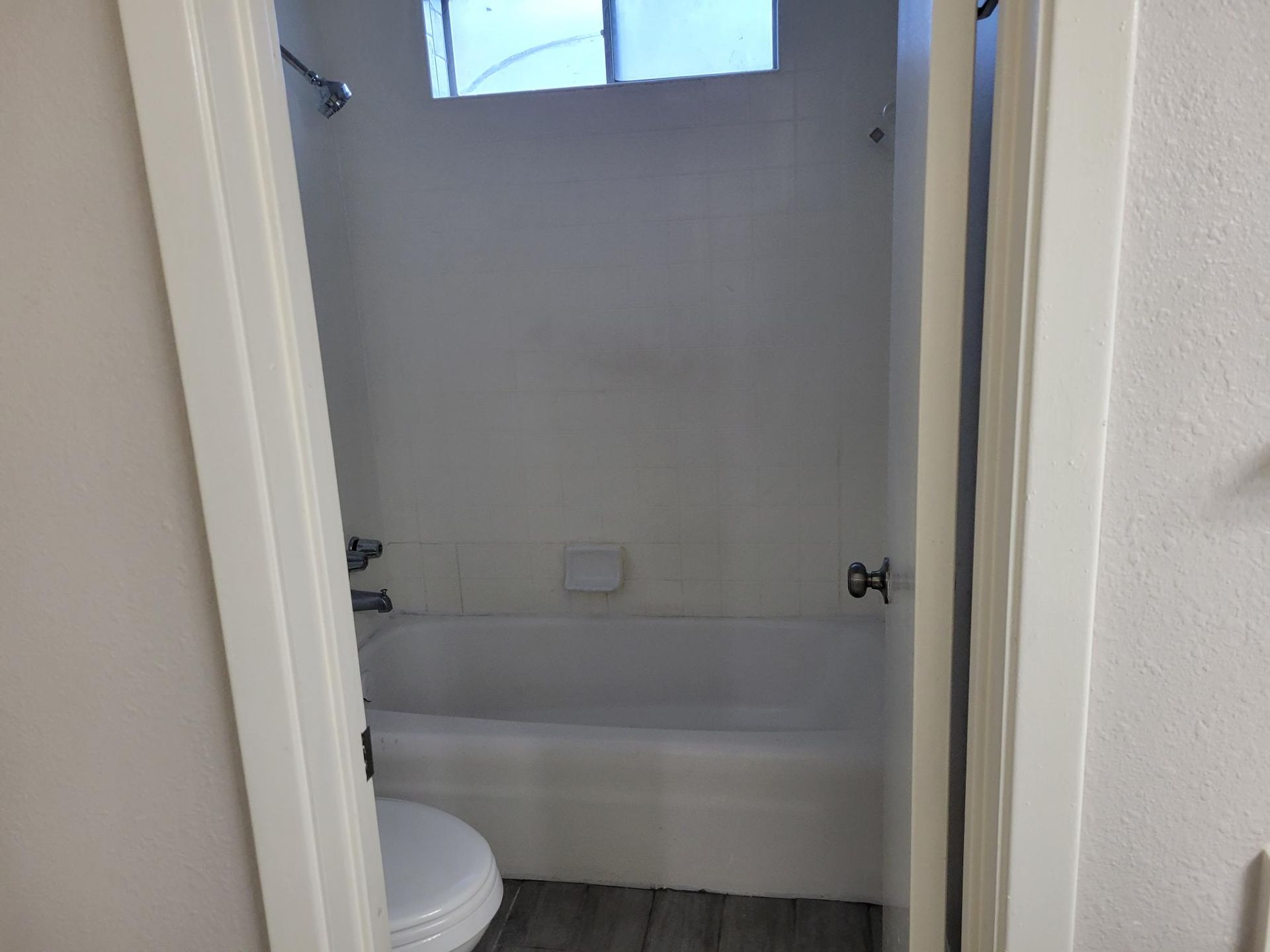 Bathroom interior, showing a toilet, bathtub, and window. White walls and door frame. Gray flooring.