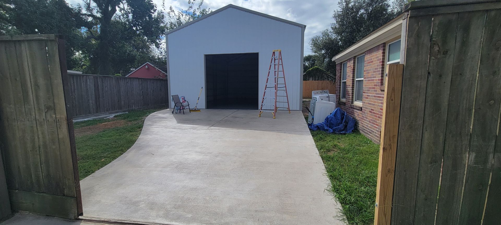 A concrete driveway leads to a white metal garage, flanked by green grass and wooden fences.