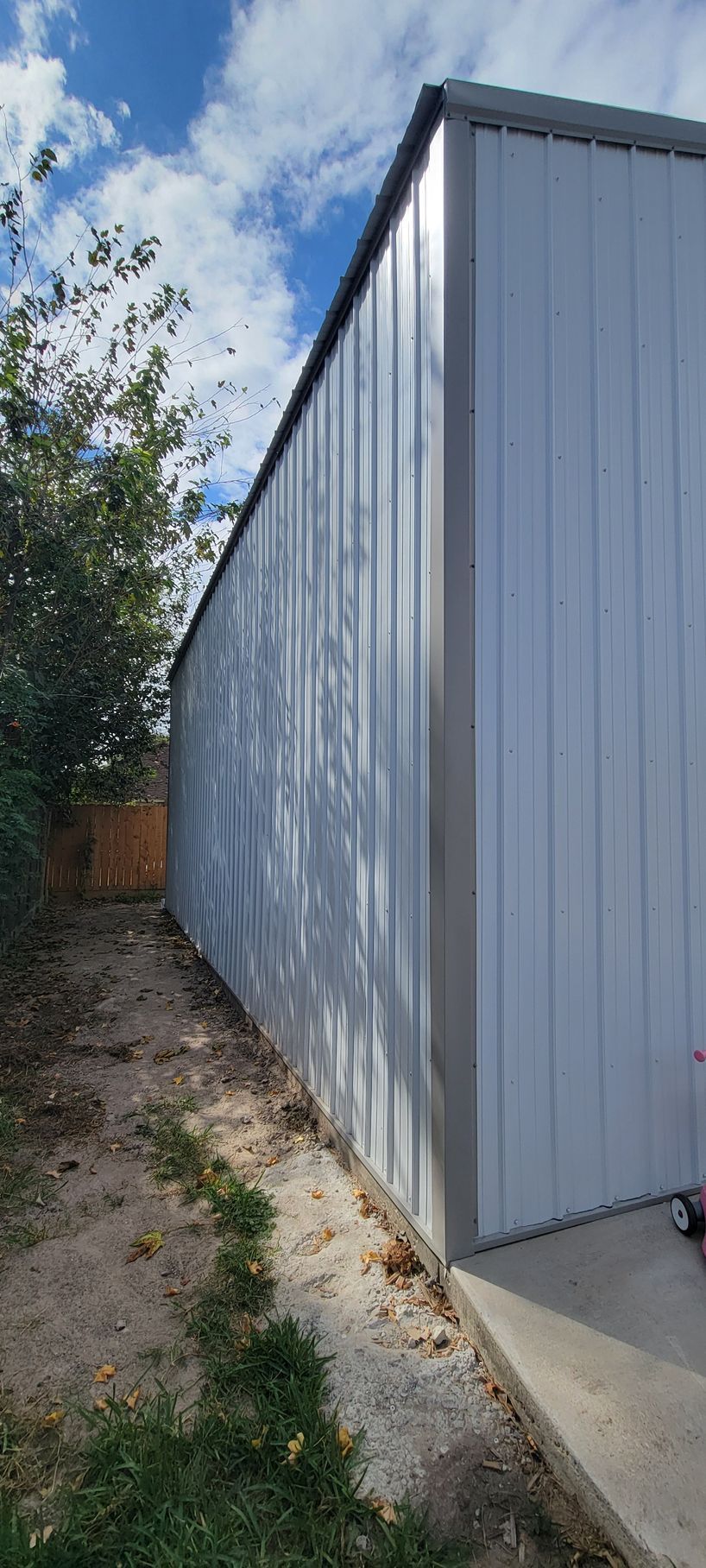 Metal shed with corrugated walls, beside a dirt and grass area. Blue sky with clouds in the background.