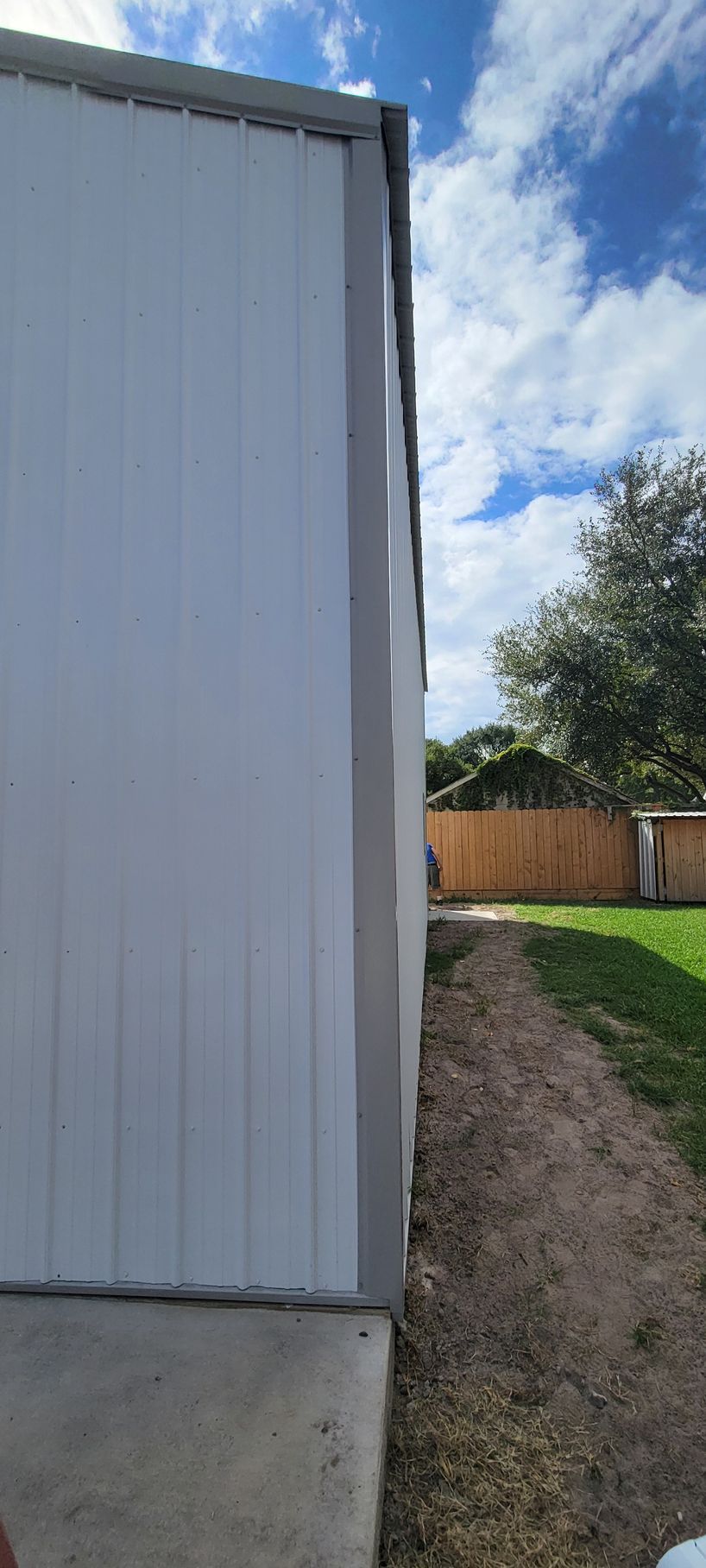 Side view of a white building with a gray trim next to a dirt path and green lawn under a blue sky.