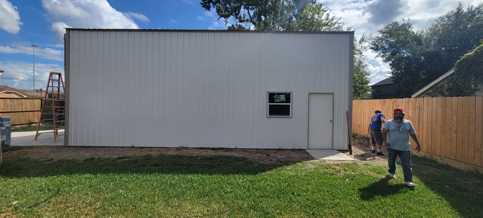 Two people walk in a grassy yard beside a metal building, a wooden fence, and a door.