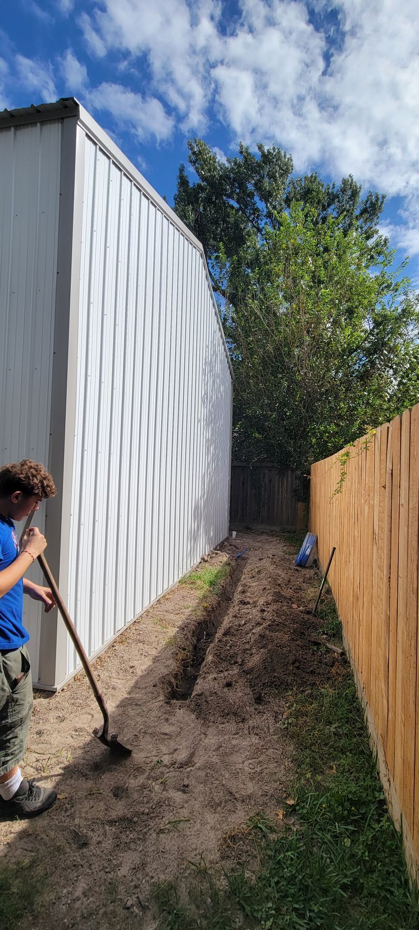 Person raking a garden bed between a white building and a wooden fence, under a partly cloudy sky.