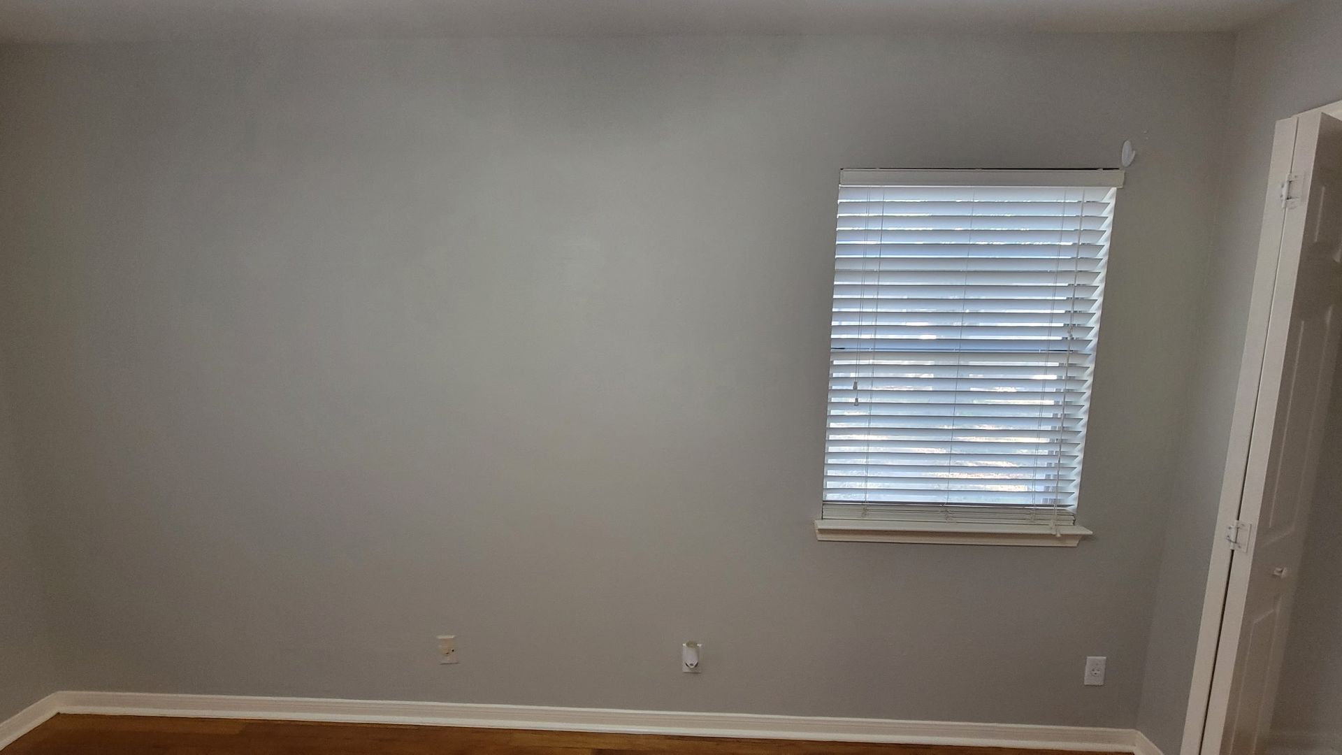 Gray room with window and closed blinds. Light shines in from the window. Brown floor.
