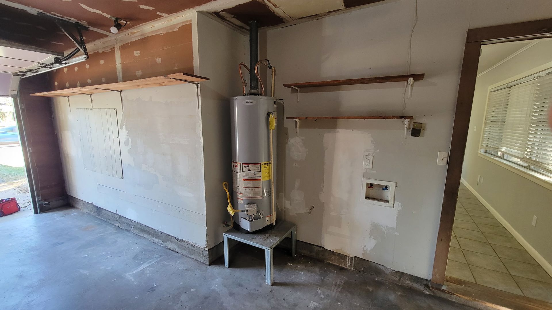 Garage interior with water heater, concrete floor, exposed wall studs, and open door to a room.