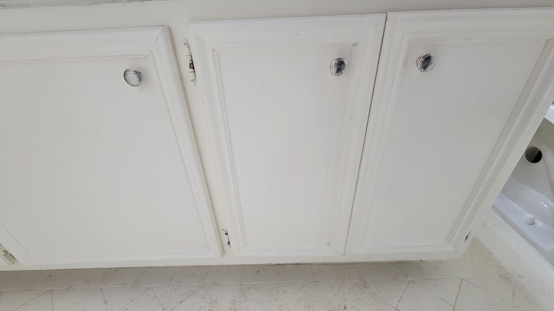 White bathroom cabinets with cracks, silver knobs, and a glimpse of a white sink.