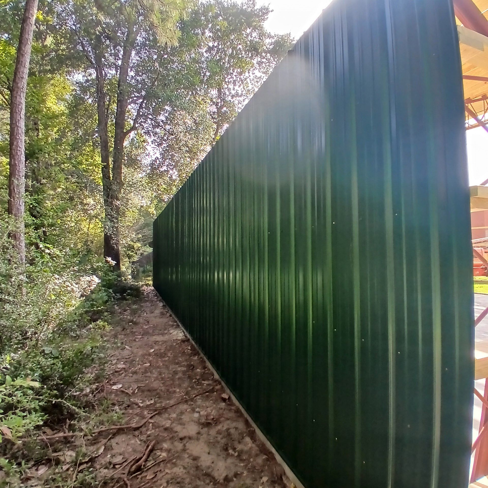 Green metal fence alongside a dirt path, with trees in the background. Sunny day.