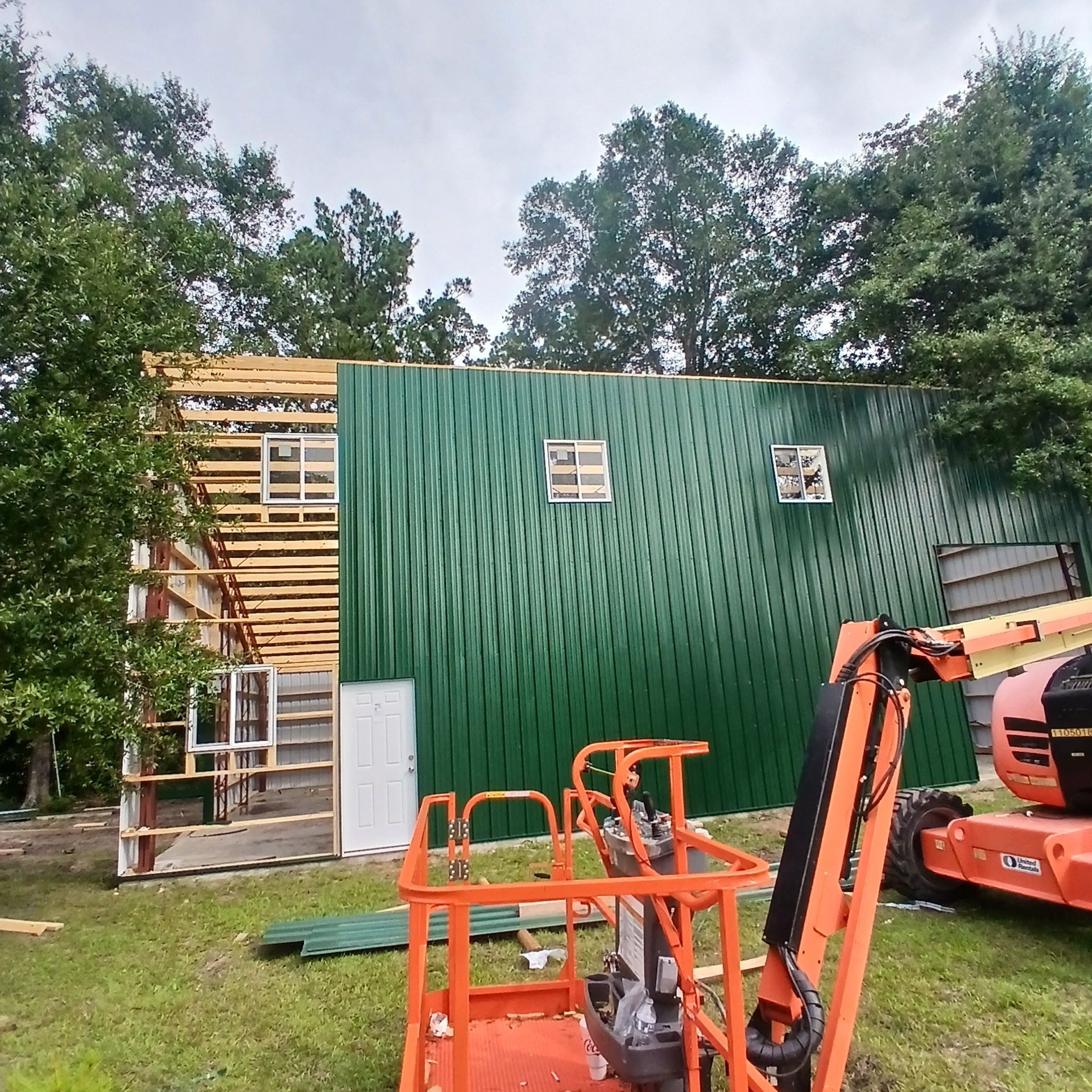 Construction site: green corrugated metal siding, lift platform, unfinished structure with exposed wood framing.