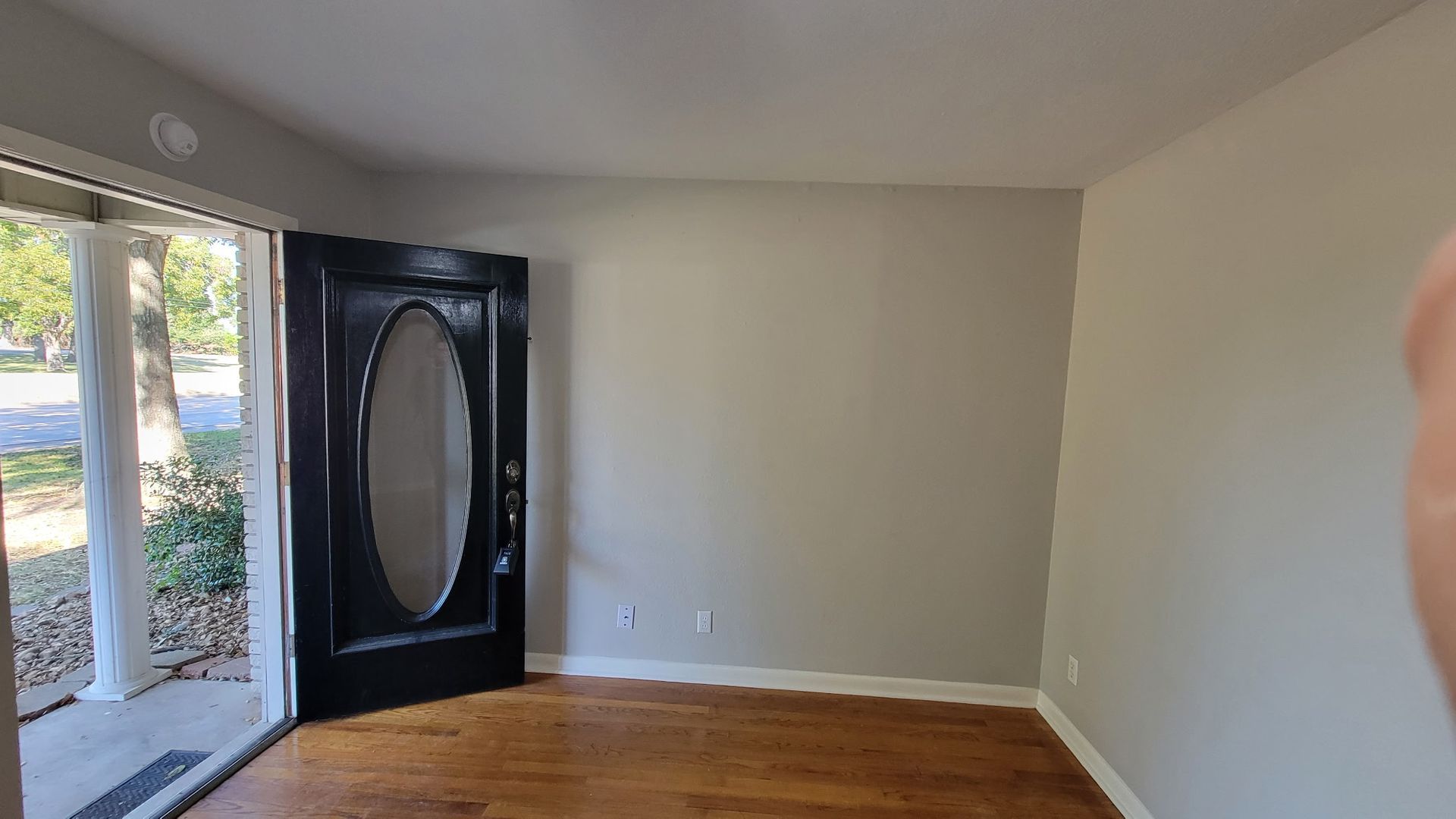 Empty room with a dark door with an oval window, facing an open doorway, hardwood floors, and light gray walls.