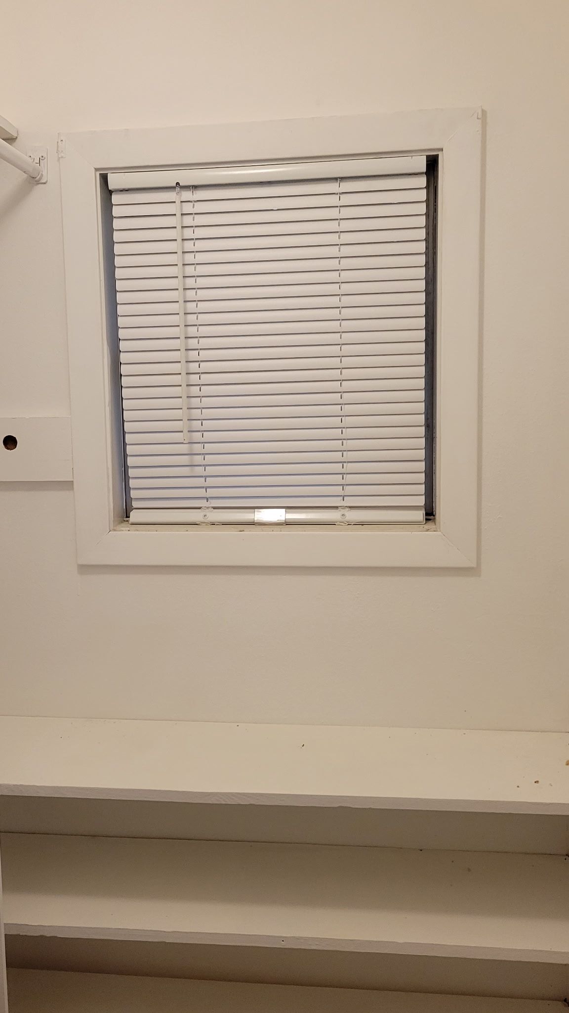 White square window with closed white blinds, set in a white wall with a white shelf below.