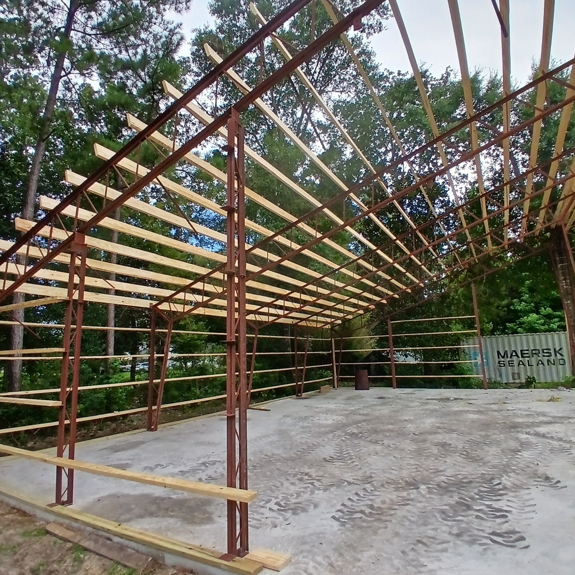 Steel and wood framing of a partially constructed building. Concrete floor. Outdoors, trees in the background.
