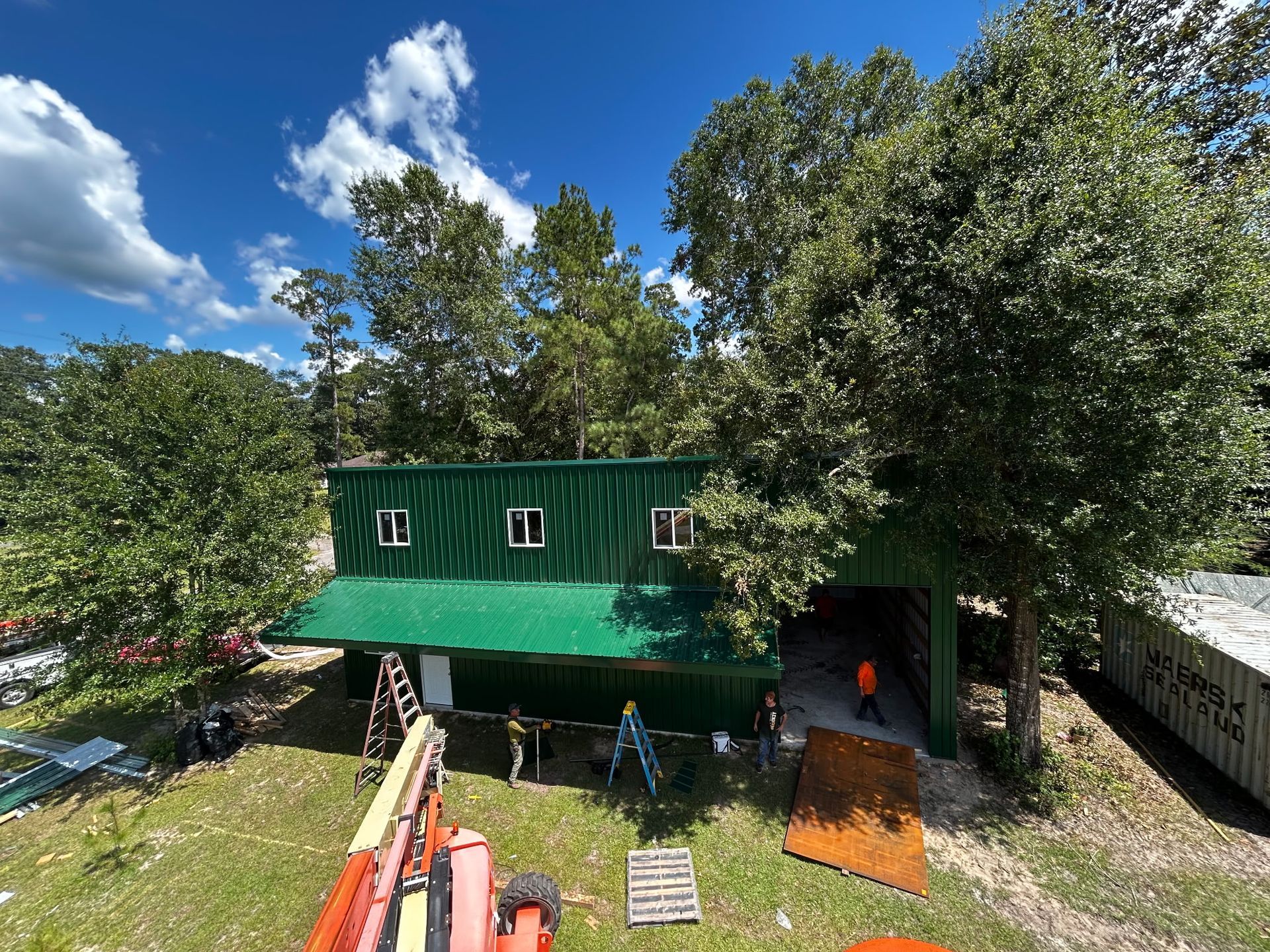 Green building with white-framed windows, surrounded by trees. Workers near a ramp and equipment outside.