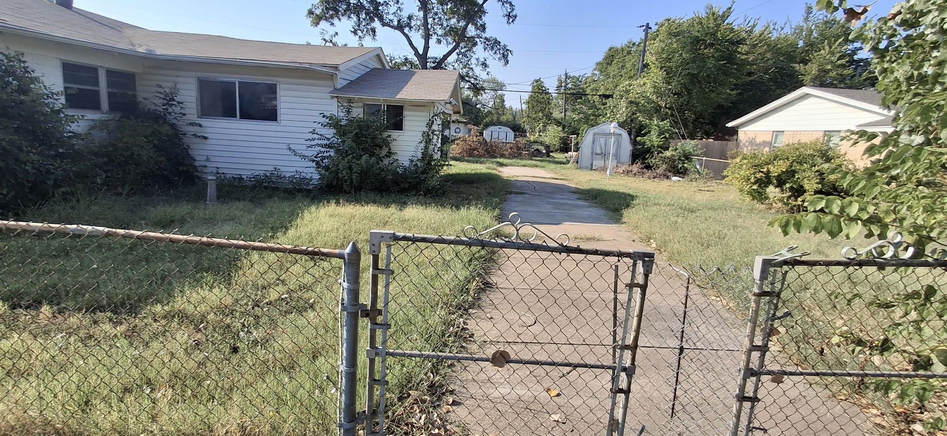 House with overgrown yard and chain-link gate. Driveway leads to a shed.