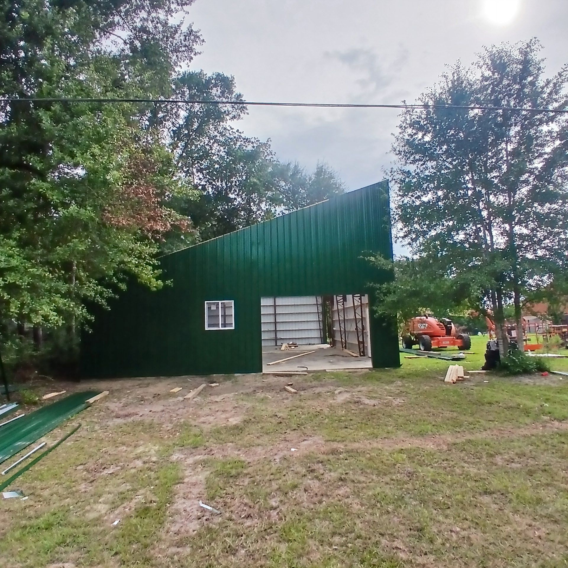 Green metal building under construction in a grassy area, trees in the background, overcast day.
