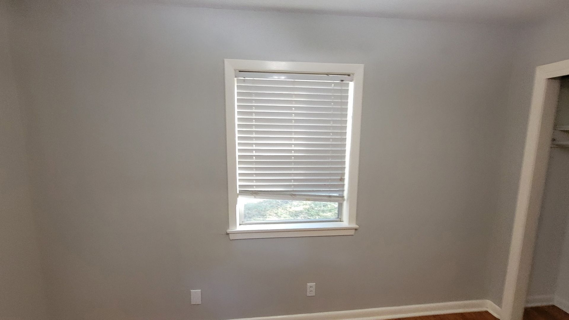 Empty room with grey walls, window with blinds, and wooden floor.