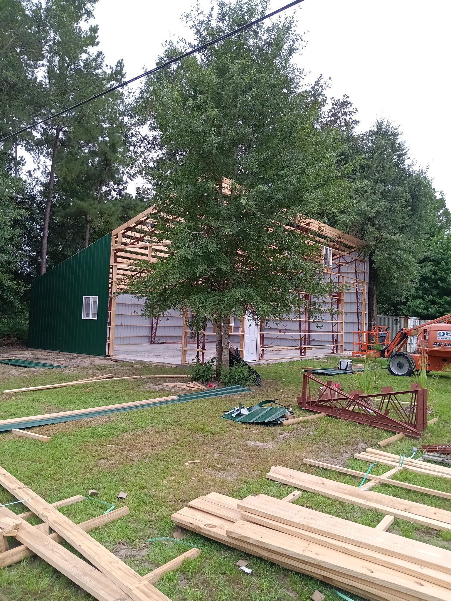Construction of a building with exposed framing, partial green siding, and a large tree in front.