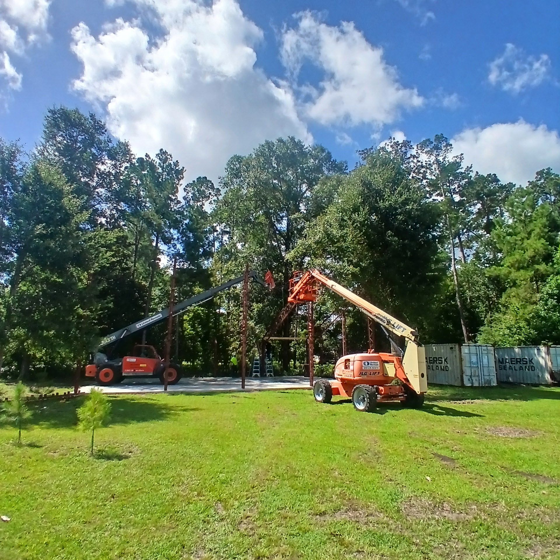 Two orange lift machines operating near trees and a building frame on a grassy area under a blue sky.