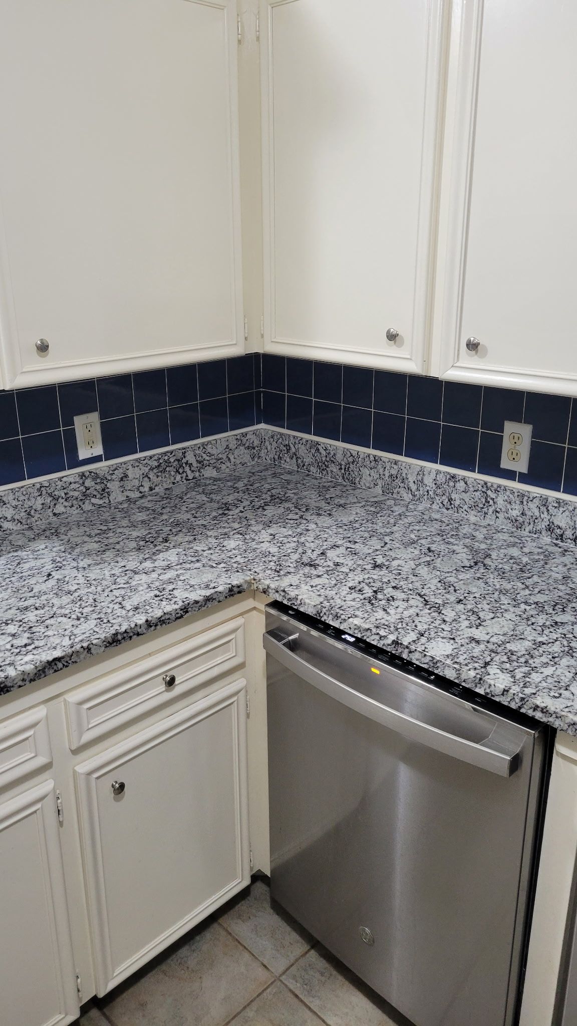 White kitchen cabinets with granite countertop, stainless steel dishwasher. Dark blue backsplash.