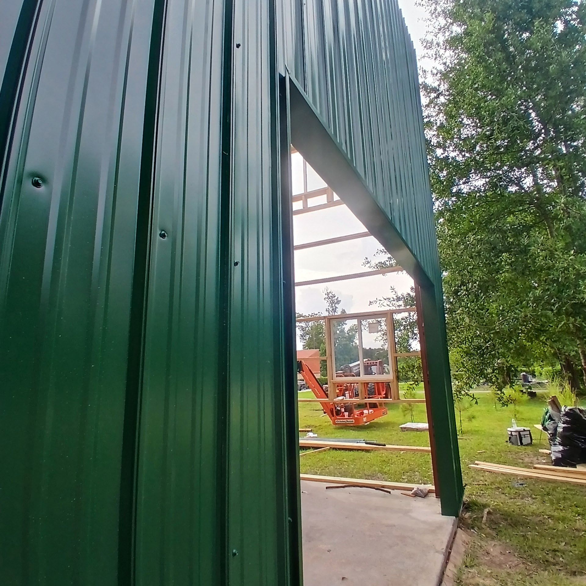 Green metal building under construction, doorway opening onto frame, grassy yard, orange machinery.