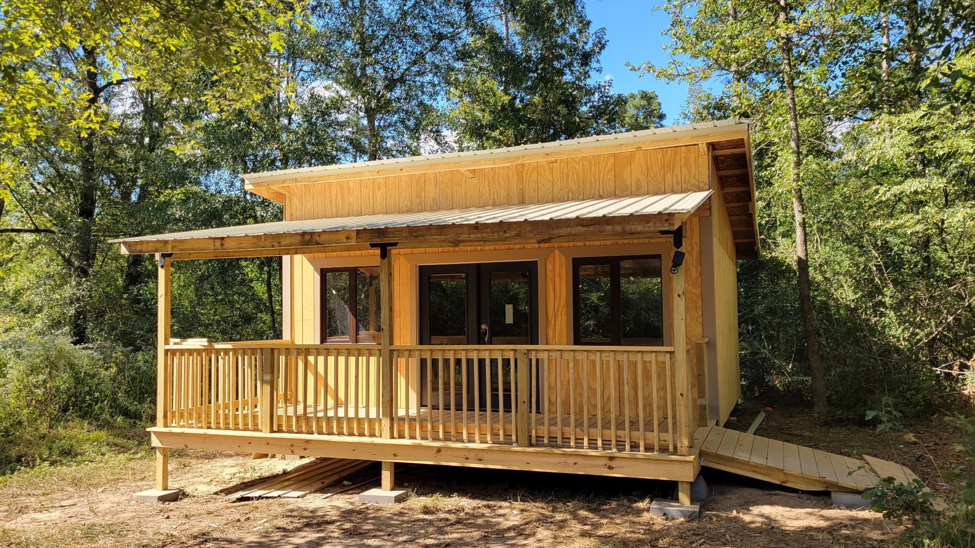 Wooden cabin with porch and ramp in a wooded area, blue sky.