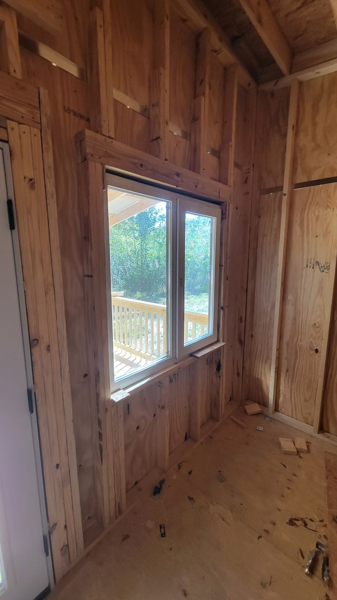 Interior view of a room under construction with a framed window overlooking a deck and trees. Wooden beams and plywood walls.