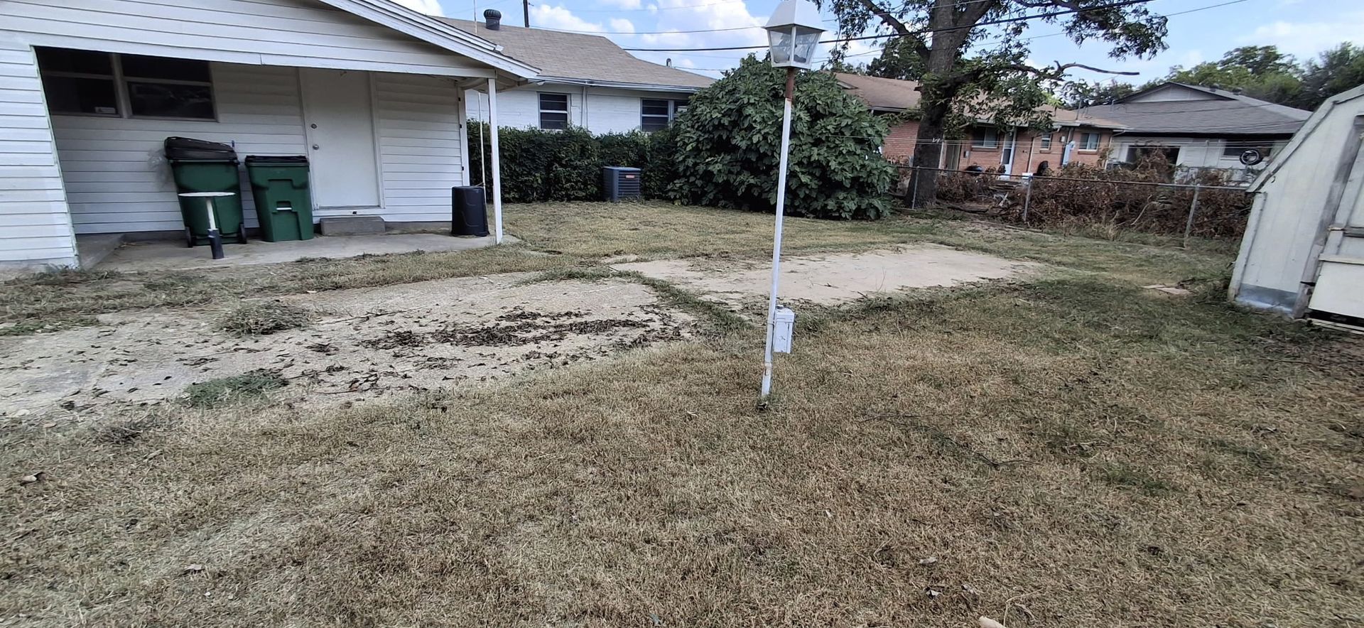 Backyard with dry grass, two green trash cans, a white light pole, and a small trailer.