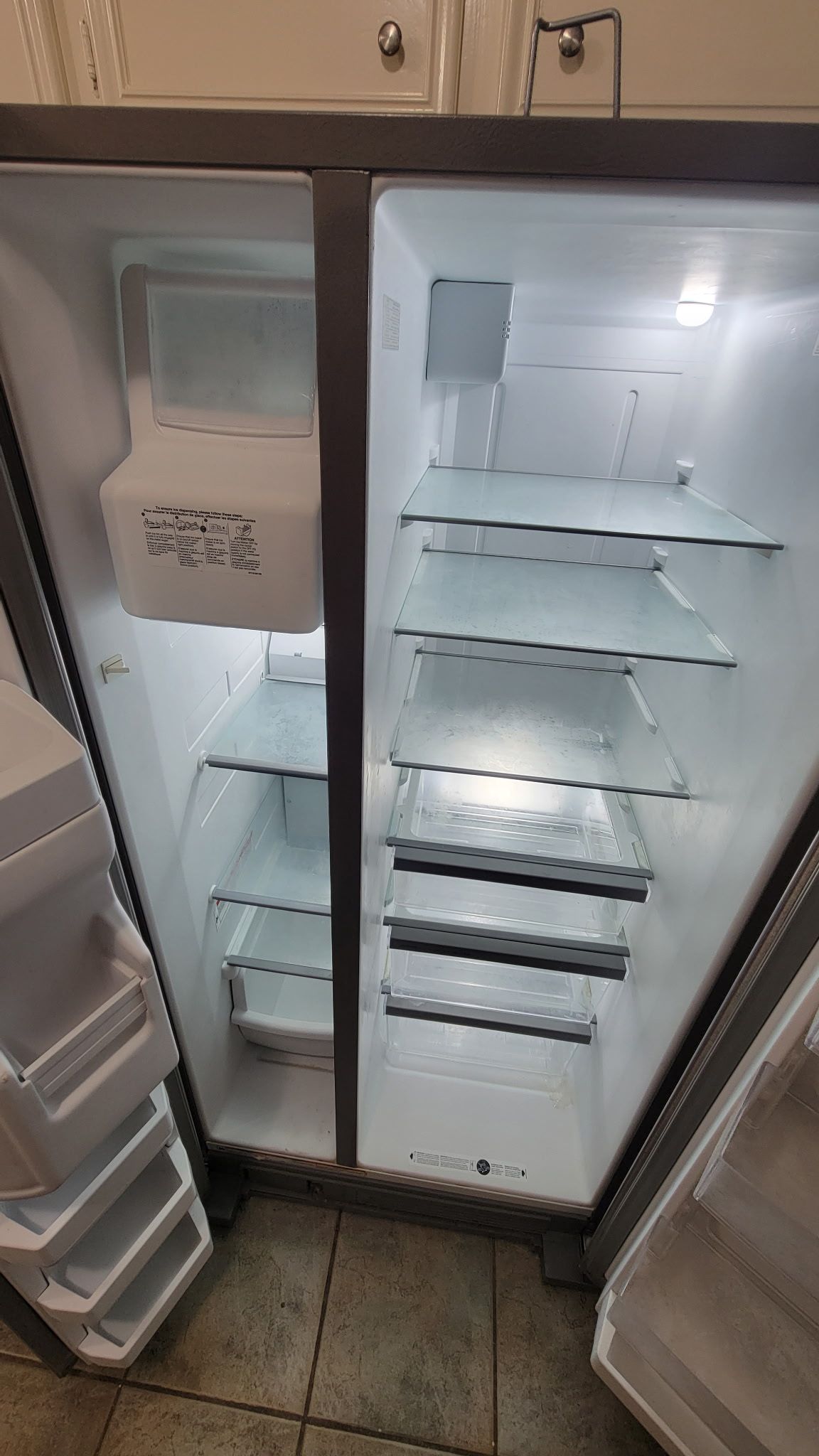 Open, empty, side-by-side refrigerator. Interior lit, with glass shelves and ice maker on the left door.