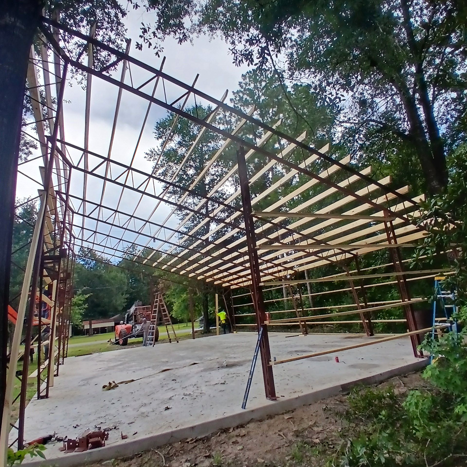 Construction of a metal framed building with wooden beams. Concrete foundation, trees, and sky in the background.