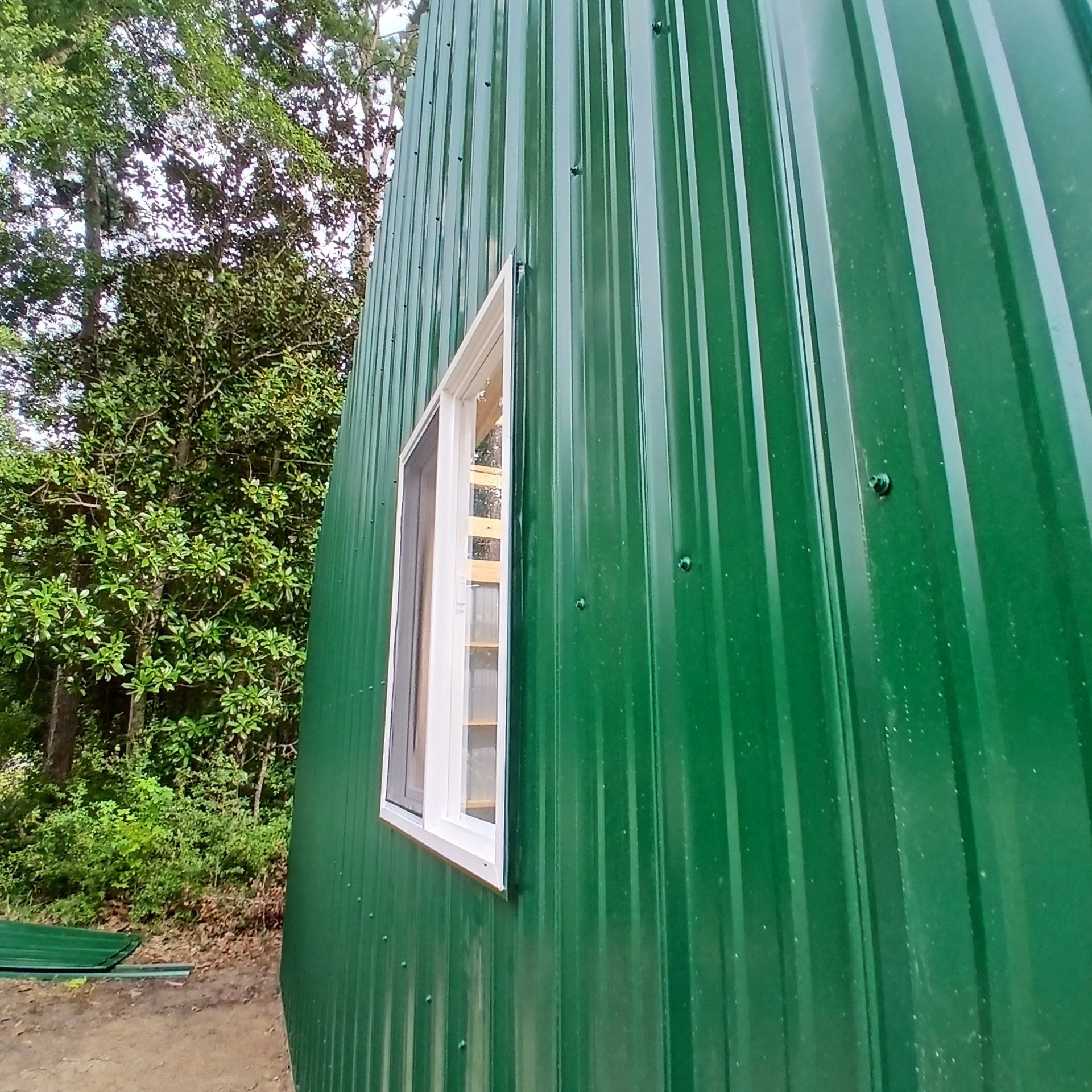 Green corrugated metal building with a white window, trees in the background.