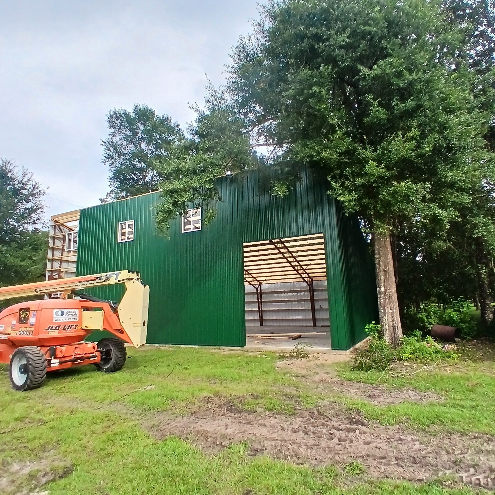 Green metal barn under construction with an orange lift, near trees.