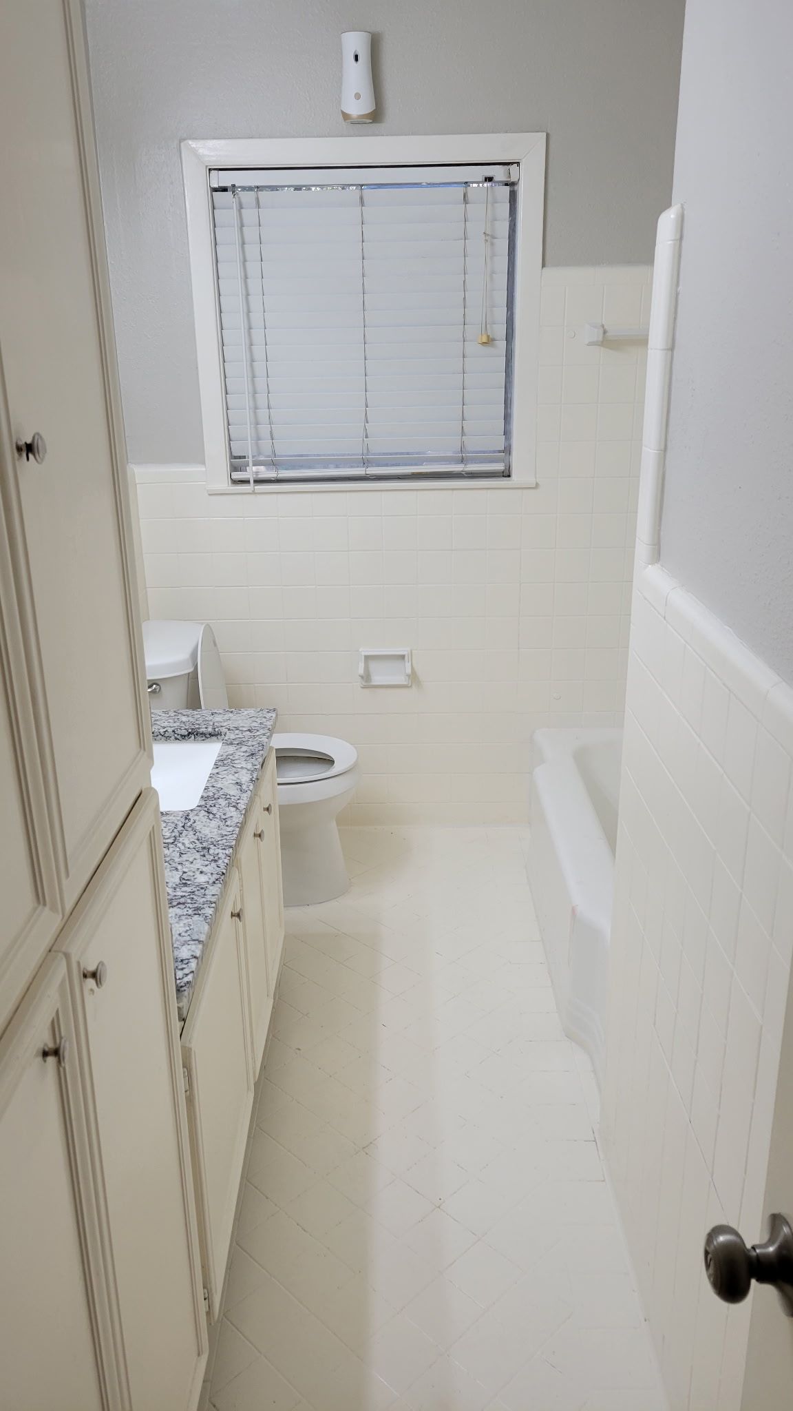 Narrow bathroom with white tile walls and floor, vanity, toilet, and bathtub.