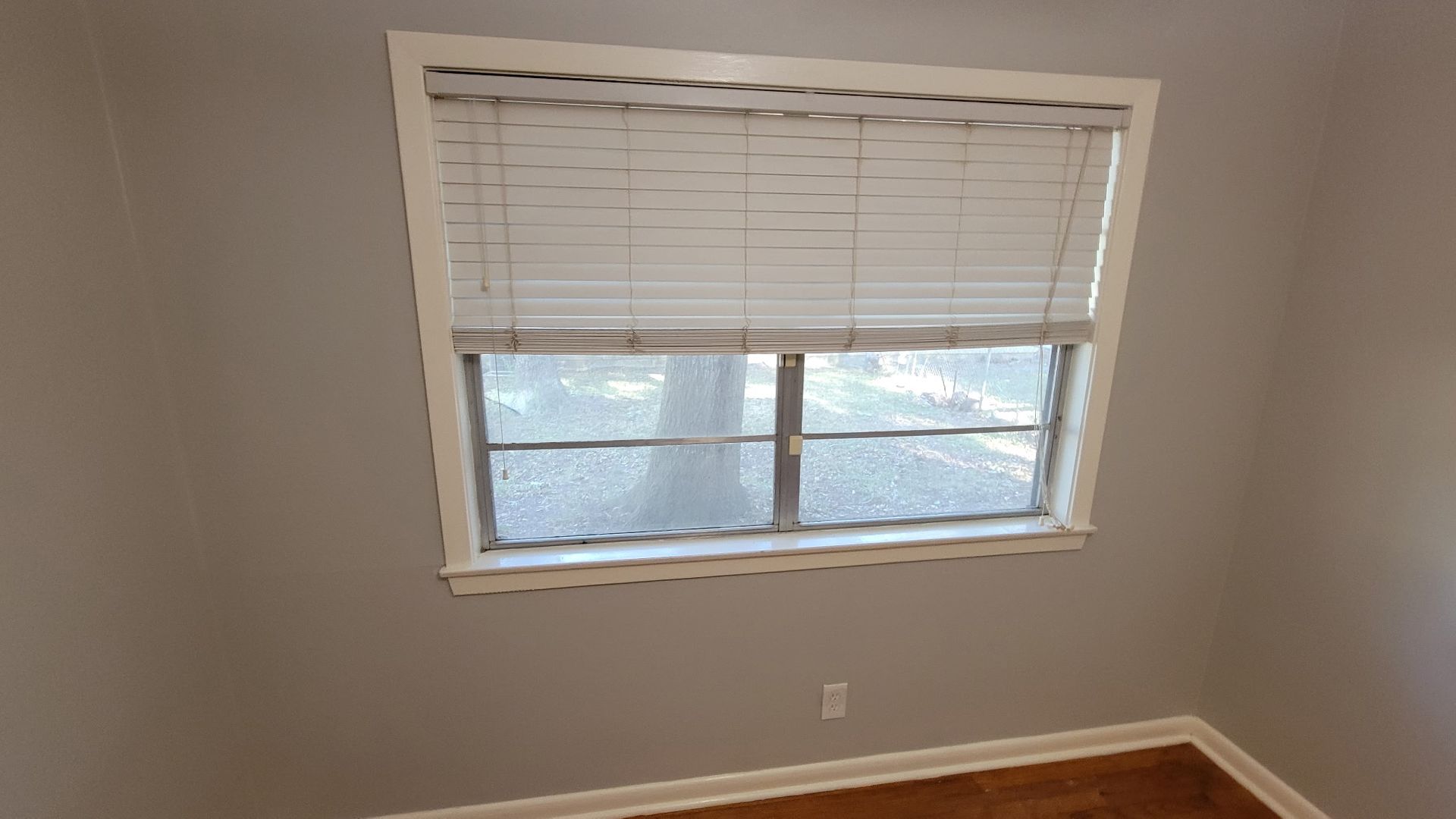 Window with white blinds and frame, gray walls, and brown flooring.