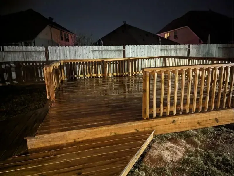 Wooden deck, partially lit, in a backyard at night, with a fence and houses in the background.