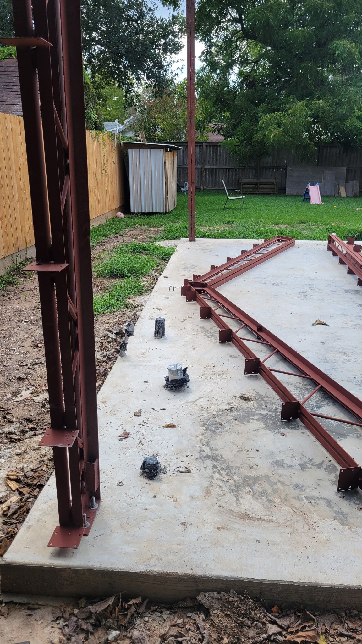 Construction site: concrete pad with metal beams, support posts, and small shed in a backyard setting.