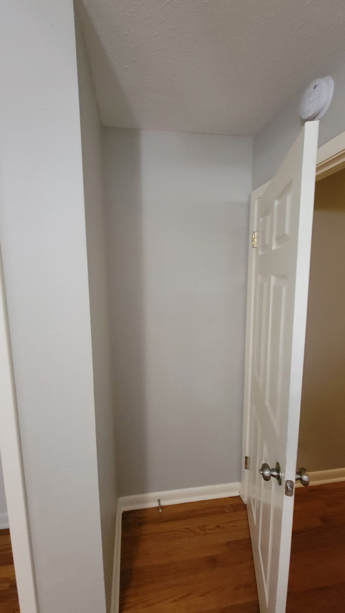 A partially open white door in a room with light gray walls and wood flooring. A smoke detector is visible.