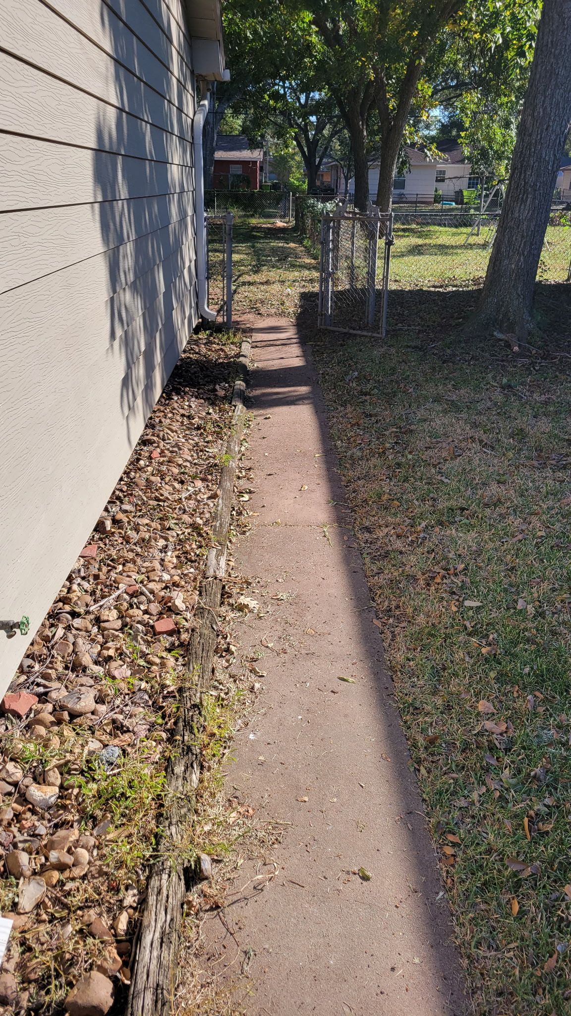A narrow concrete walkway between a house and a grassy yard, partially covered in leaves.