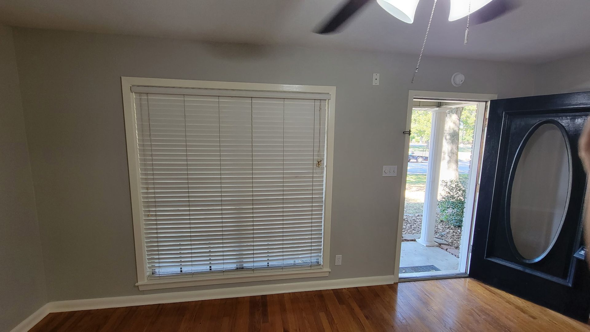 A room with a window with blinds, a black door, and hardwood floors.