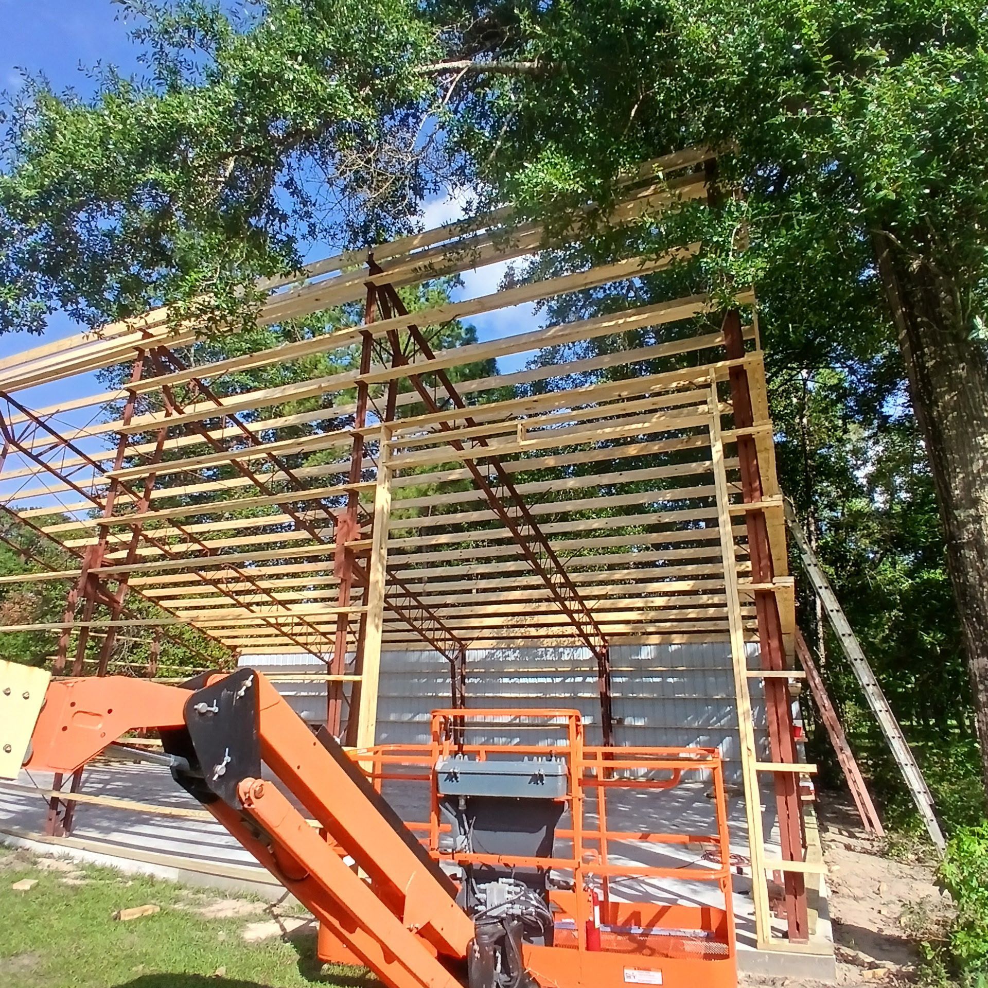 Construction of a metal framed building with wooden beams. Orange lift is in front. Trees surround.